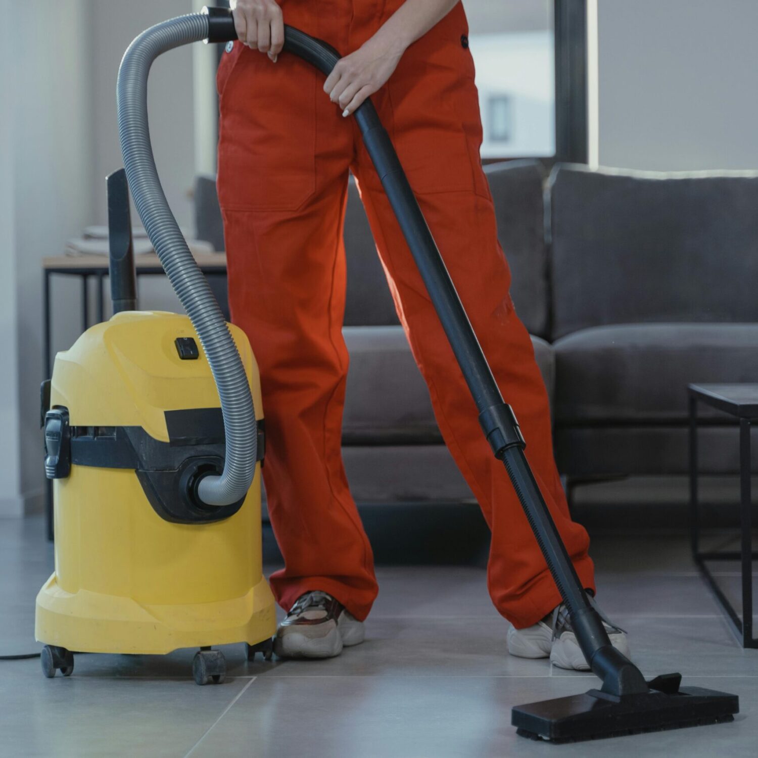A professional cleaner in red coveralls vacuums a contemporary living room with a yellow vacuum cleaner.